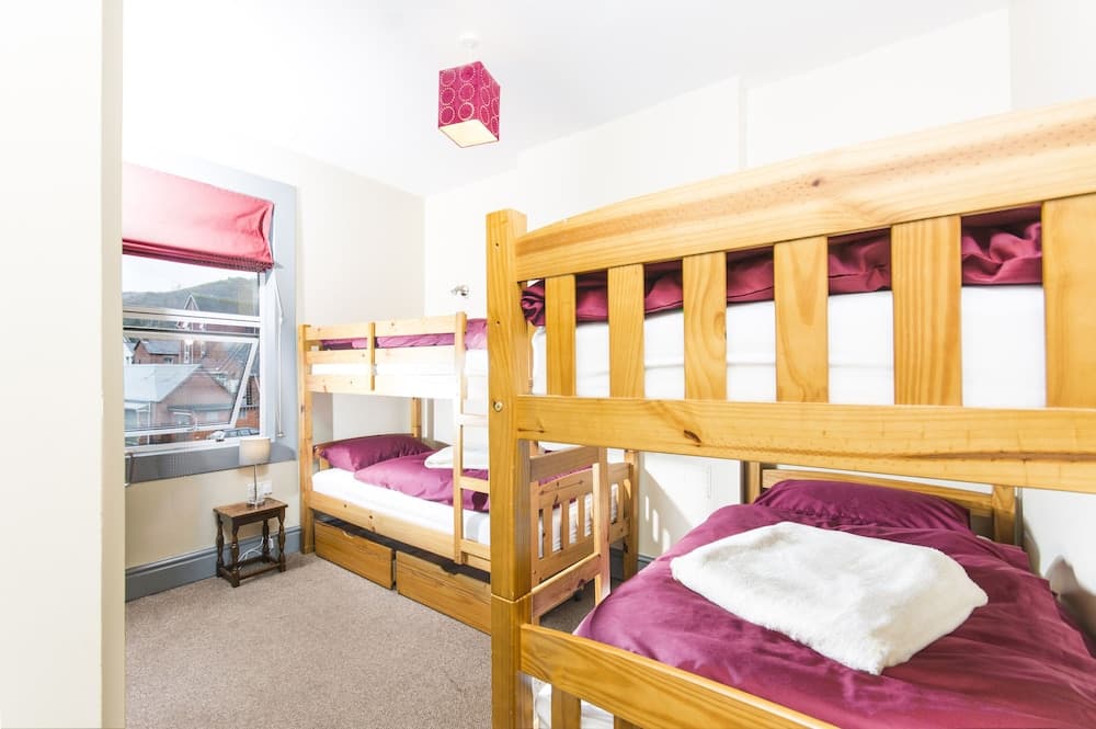 A cozy hostel room with wooden bunk beds adorned in maroon bedding, a small bedside table, and a window view of rooftops, ideal for budget travelers seeking comfort and simplicity.