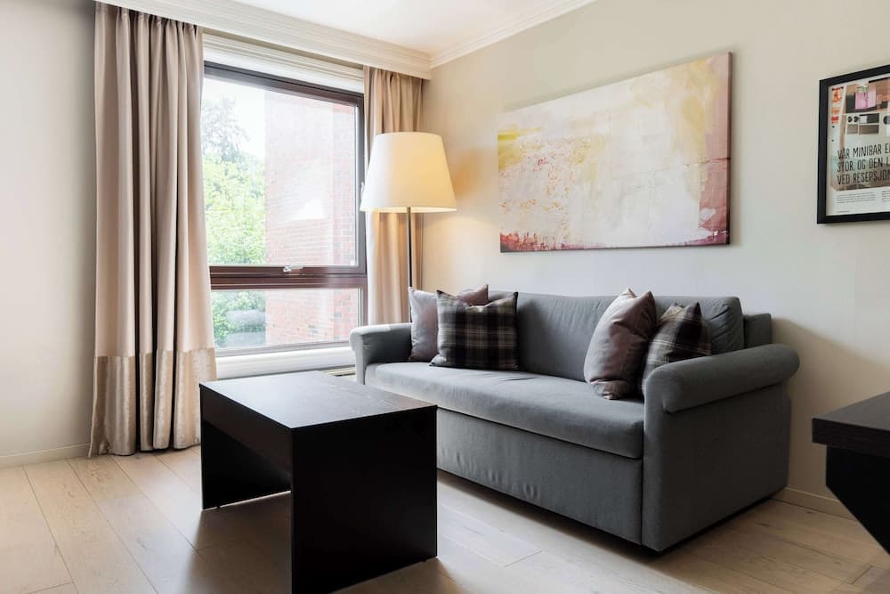 A cozy living room with a gray sofa adorned with patterned pillows, a sleek black coffee table, and large windows framed by beige curtains, creating a serene and inviting atmosphere ideal for a relaxing getaway.