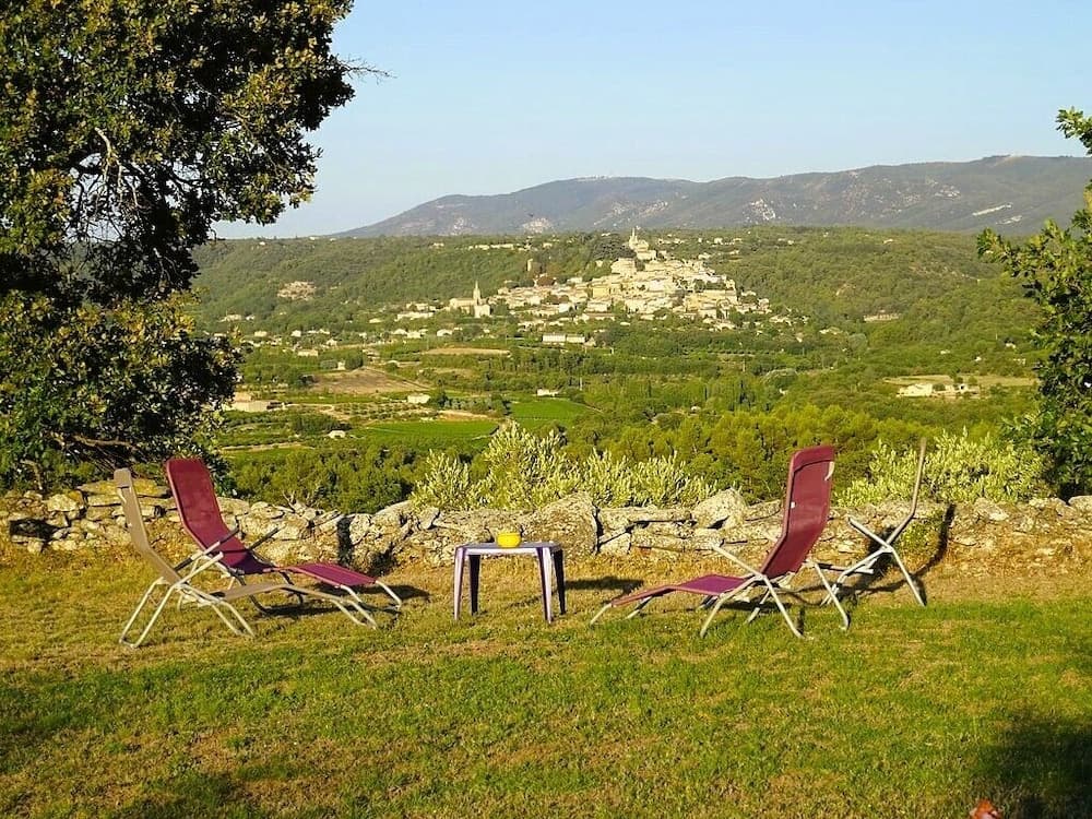 A serene outdoor seating area with lounge chairs overlooks a picturesque hillside village nestled among lush greenery and distant mountains, ideal for travelers seeking relaxation and scenic views.