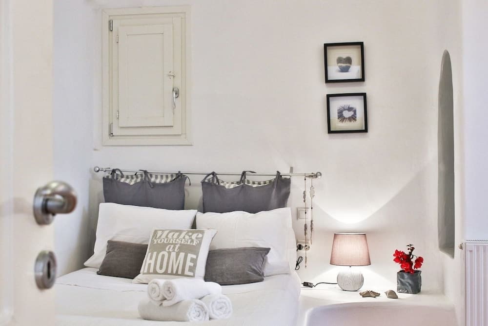A cozy bedroom with white walls, a neatly made bed adorned with "Make Yourself at HOME" pillows, soft lighting from a bedside lamp, and decorative elements like framed hearts and red flowers creates an inviting atmosphere perfect for travelers seeking comfort and relaxation.
