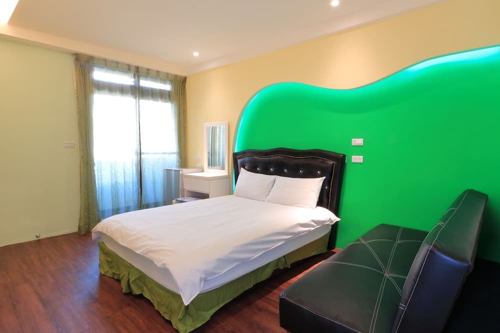 A cozy hotel room features a neatly made bed with white linens against a vibrant green accent wall, complemented by a leather bench, wooden flooring, and soft natural light filtering through sheer curtains, ideal for travelers seeking comfort and style.