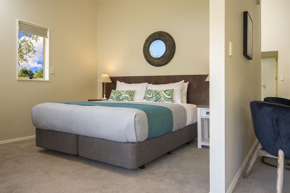 A cozy bedroom with a neatly made bed adorned with green and white pillows, a round wall mirror, and a window offering a serene outdoor view, ideal for travelers seeking relaxation in a modern vacation rental.