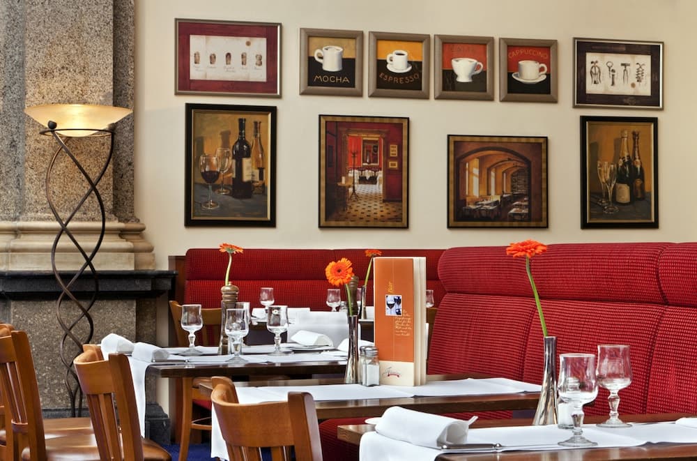 A cozy restaurant interior features red upholstered benches, framed coffee-themed artwork, elegant table settings with vibrant orange flowers, creating an inviting atmosphere ideal for a romantic dinner or business meeting.