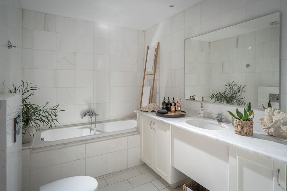 A serene, all-white bathroom features a bathtub, marble countertop with plants and decor, creating a tranquil spa-like atmosphere ideal for relaxation and wellness retreats.