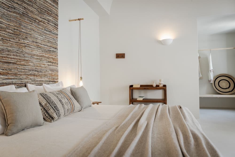 A serene minimalist bedroom features a cozy bed adorned with patterned pillows, a textured wall hanging, and soft lighting, ideal for travelers seeking relaxation and comfort.