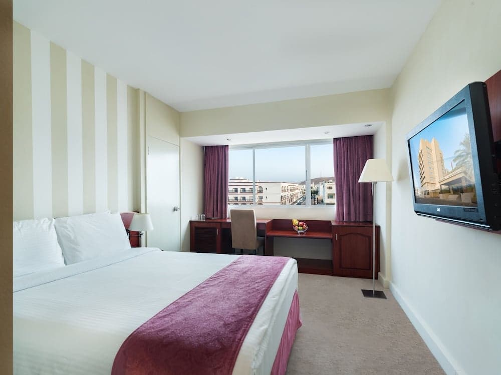 A cozy hotel room with a neatly made bed, striped walls, a desk by the window overlooking urban scenery, and a mounted TV creates an inviting atmosphere for travelers seeking comfort and convenience.