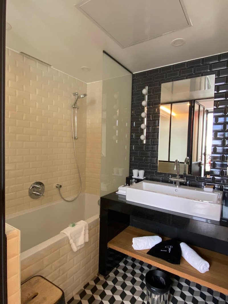 A modern bathroom at the Terrass Hotel in Paris features a stylish black-and-white tiled floor, a sleek bathtub with a shower, and a vanity with a large mirror, ideal for travelers seeking comfort and elegance during their stay.