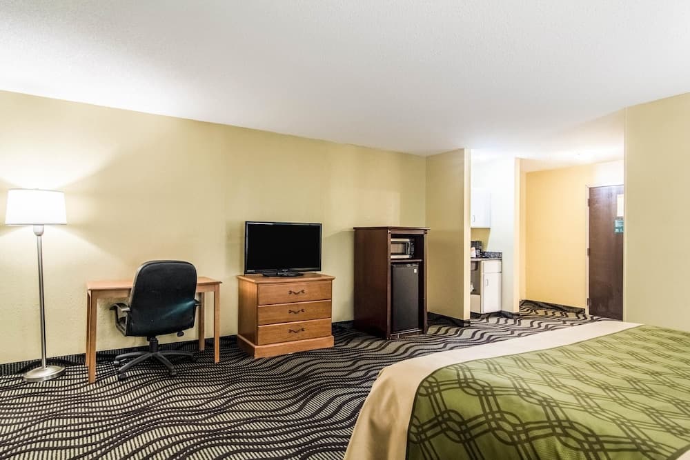 A cozy hotel room features a neatly made bed, a desk with a chair, a flat-screen TV on a wooden dresser, and a small kitchenette in the background, ideal for travelers seeking comfort and convenience.