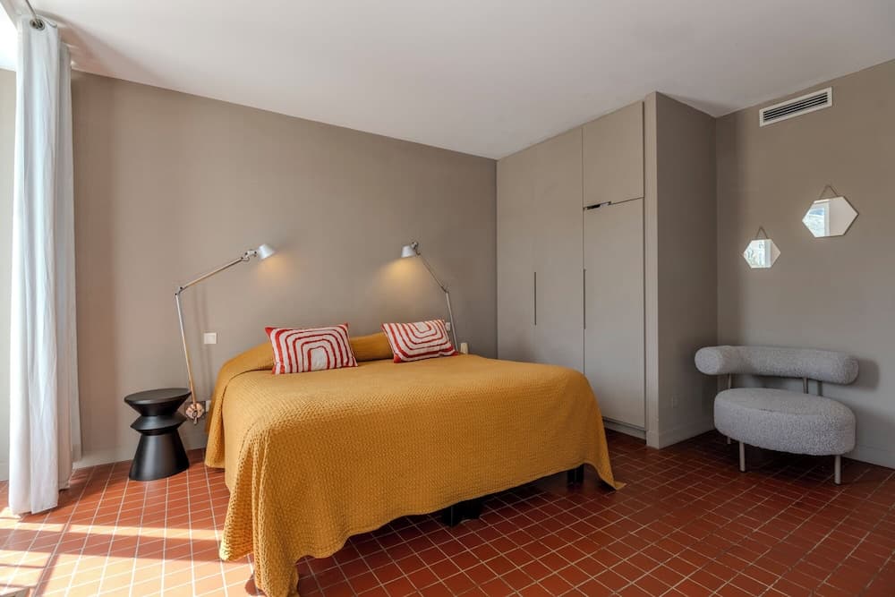 A cozy bedroom with terracotta tiled floors, a mustard yellow bedspread, modern furnishings, and geometric wall mirrors exudes a warm and inviting atmosphere, ideal for travelers seeking a stylish and comfortable retreat.