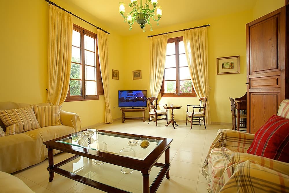 A cozy living room with yellow walls, wooden furniture, and large windows draped in cream curtains creates a warm and inviting atmosphere, perfect for travelers seeking a comfortable and charming accommodation.