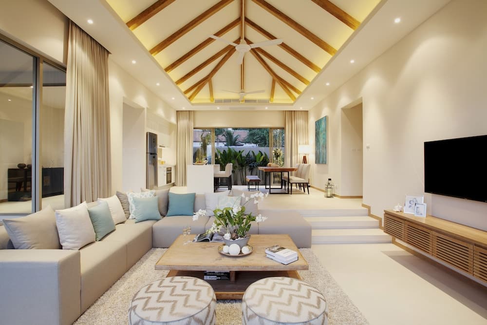 A serene tropical villa living room features a vaulted ceiling with exposed wooden beams, plush seating, and large windows opening to lush greenery, ideal for travelers seeking a luxurious and relaxing retreat.