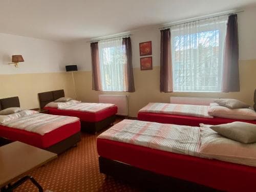 A cozy dormitory-style room with four single beds adorned in red and white bedding, featuring large windows with curtains, framed artwork on the walls, and a warm ambiance ideal for budget travelers seeking comfortable group accommodations.