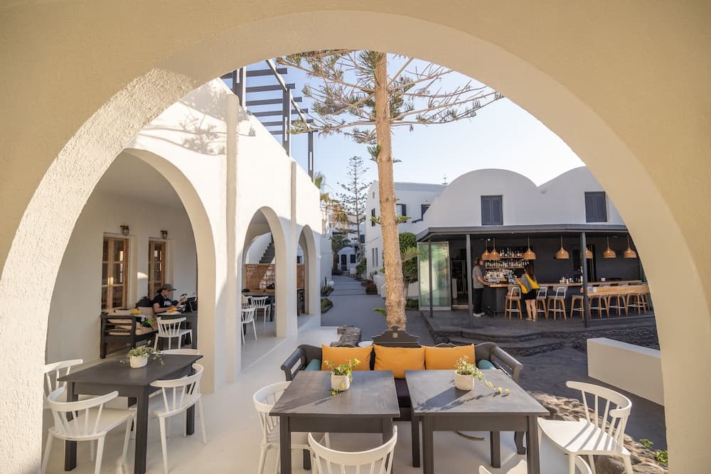 A serene outdoor café framed by white arches features cozy seating, vibrant orange cushions, and lush greenery, ideal for travelers seeking a tranquil dining experience in a Mediterranean-style setting.