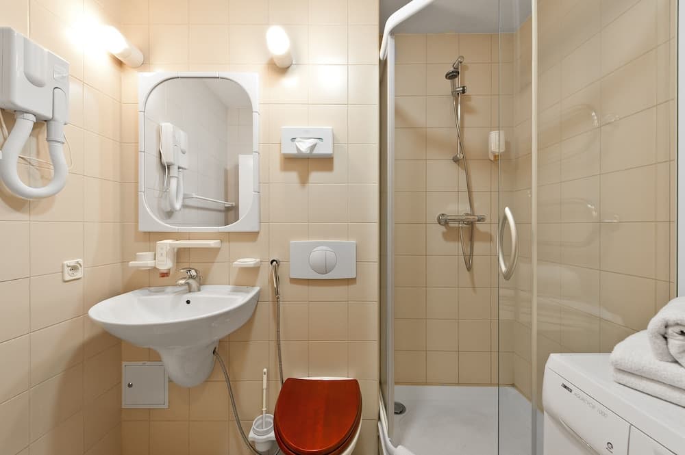 A cozy hotel bathroom features beige tiled walls, a modern sink with a mirror, a red toilet seat, a glass-enclosed shower, and a wall-mounted hairdryer, ideal for travelers seeking comfort and convenience.