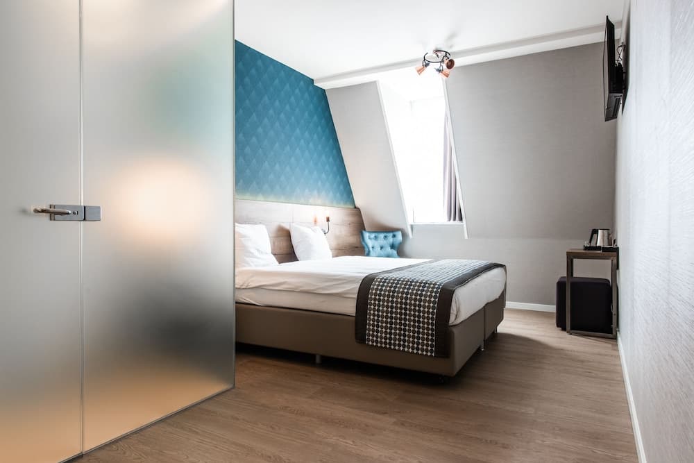 A cozy modern bedroom with a sleek bed, geometric blue wall art, and natural light from a slanted window creates a serene atmosphere, ideal for travelers seeking comfort in boutique hotels.
