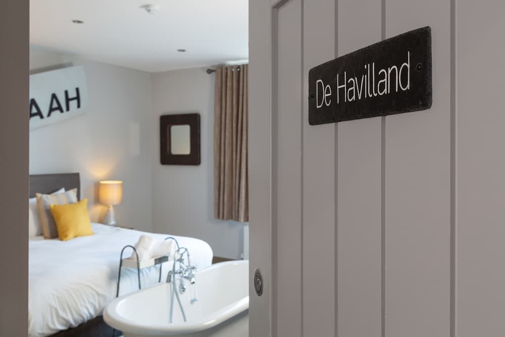 A cozy hotel room with a freestanding bathtub, neatly made bed adorned with yellow pillows, and a "De Havilland" door sign creates an inviting atmosphere ideal for travelers seeking relaxation and comfort.