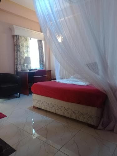 A cozy hotel room features a bed draped in white mosquito netting, red bedding, marble flooring, and a window with curtains, creating a serene and inviting atmosphere ideal for travelers seeking comfort and relaxation.
