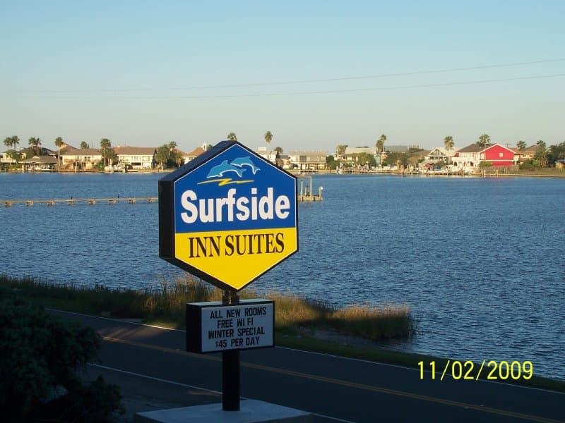 A serene waterfront scene features the Surfside Inn Suites sign by a calm bayou, ideal for travelers seeking coastal relaxation with amenities like free Wi-Fi and winter specials.