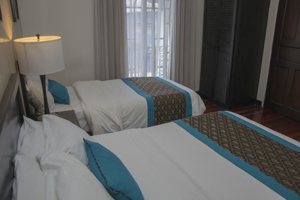 A cozy hotel room features two neatly made beds with turquoise accents, soft lighting from a bedside lamp, and a serene view through sheer curtains, ideal for travelers seeking comfort and relaxation.