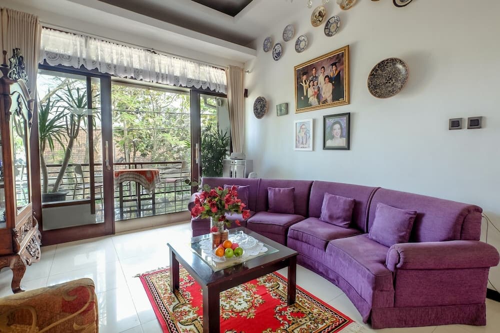 A cozy living room with a plush purple sofa, vibrant red rug, and framed family photos exudes warmth and comfort, ideal for travelers seeking a homely retreat with a view of lush greenery outside.