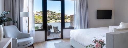 A serene hotel room with a cozy bed, modern furnishings, and a scenic view of lush hills through large glass doors, ideal for travelers seeking relaxation and comfort.