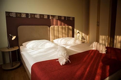 A cozy hotel room bathed in warm sunlight features a neatly made bed with white linens, a red blanket, and decorative pillows, creating an inviting atmosphere ideal for travelers seeking comfort and relaxation.