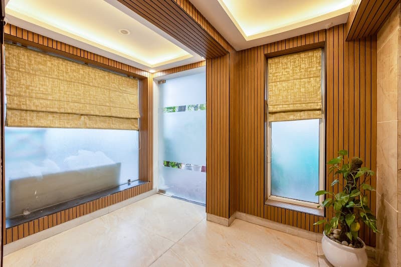A cozy indoor lounge at a hotel in Noida, featuring warm wooden paneling, frosted glass windows with golden blinds, soft lighting, and a potted plant, creating an inviting atmosphere ideal for travelers seeking relaxation.