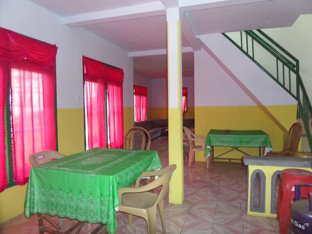 A cozy indoor dining area with vibrant green tables, pink curtains, and a staircase, ideal for travelers seeking a homely and affordable accommodation experience.
