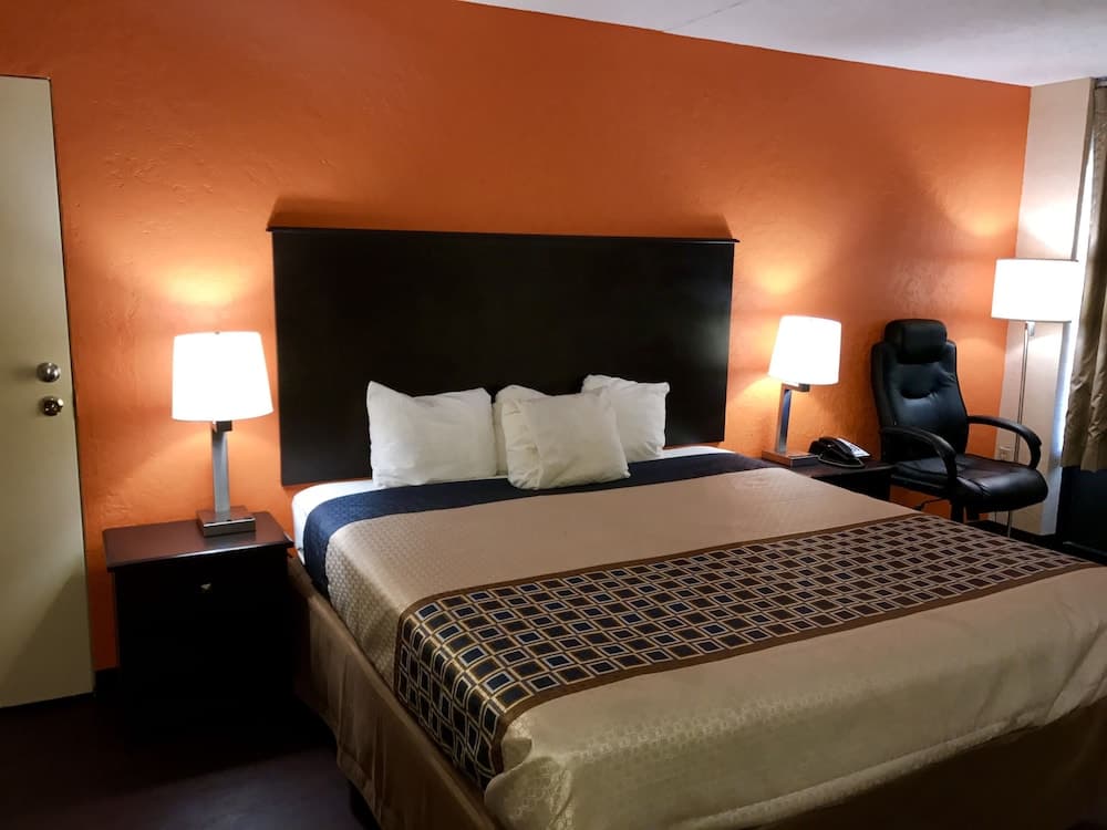 A cozy hotel room features an inviting bed with white pillows and patterned bedding against a warm orange wall, complemented by bedside lamps and a comfortable chair, ideal for travelers seeking relaxation.