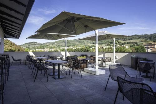 A serene rooftop terrace with tables, chairs, and large umbrellas overlooks lush green hills under a clear blue sky, ideal for travelers seeking relaxation and scenic views.
