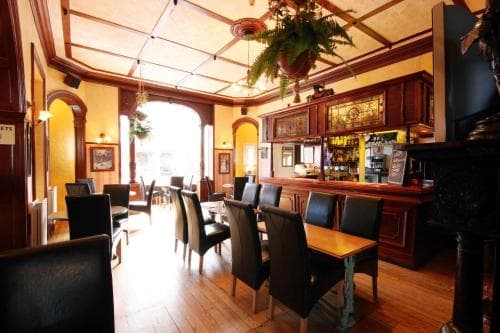 A cozy, warmly lit pub features wooden interiors, hanging plants, and inviting tables, perfect for travelers seeking a charming spot to unwind and enjoy a drink.