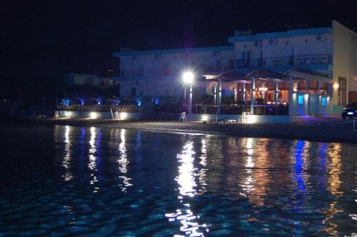 A serene nighttime beach scene illuminated by vibrant lights reflecting on the water, featuring a cozy waterfront restaurant with outdoor seating, ideal for travelers seeking a tranquil evening dining experience.