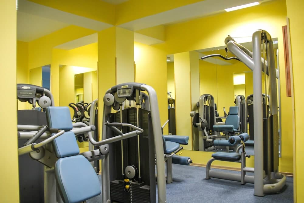 A brightly lit gym with yellow walls and blue cushioned exercise machines reflected in mirrors creates an inviting atmosphere for fitness enthusiasts seeking a well-equipped workout space.