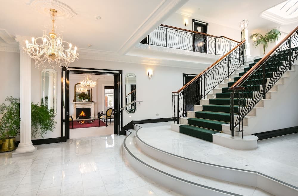 A luxurious mansion foyer features a grand staircase with green carpeting, marble floors, a chandelier, and elegant decor, ideal for high-end real estate listings or luxury travel inspiration.