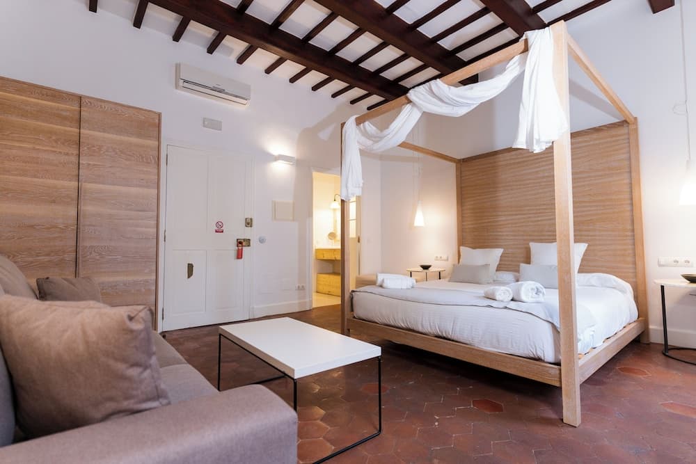A cozy bedroom with rustic wooden beams, terracotta flooring, and a four-poster bed draped in white fabric creates an inviting atmosphere ideal for travelers seeking a charming and comfortable retreat.