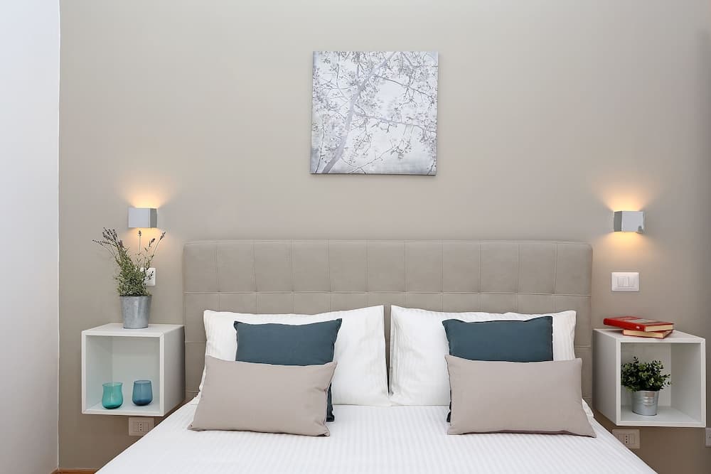 A serene hotel bedroom features a neatly made bed with plush pillows, minimalist nightstands holding plants and books, soft wall-mounted lights, and a calming tree branch artwork, ideal for travelers seeking comfort and tranquility.