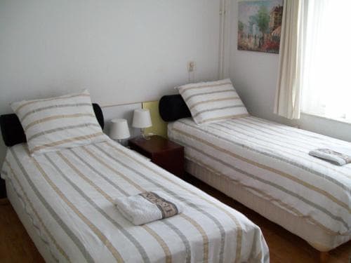 A cozy hotel room features two neatly made single beds with striped linens, a small bedside table with lamps, and a bright window, ideal for travelers seeking a comfortable and simple accommodation.