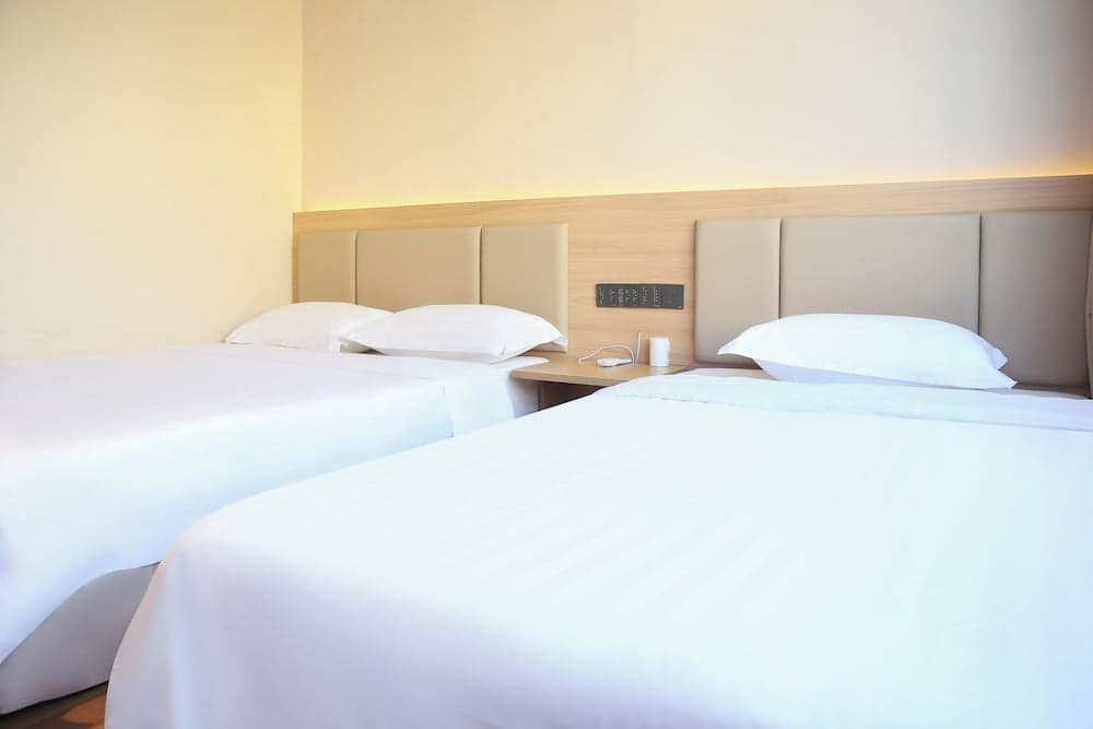 A cozy hotel room features two neatly made beds with white linens, a wooden bedside table with a lamp and remote control, set against soft yellow walls, ideal for travelers seeking comfort and simplicity.
