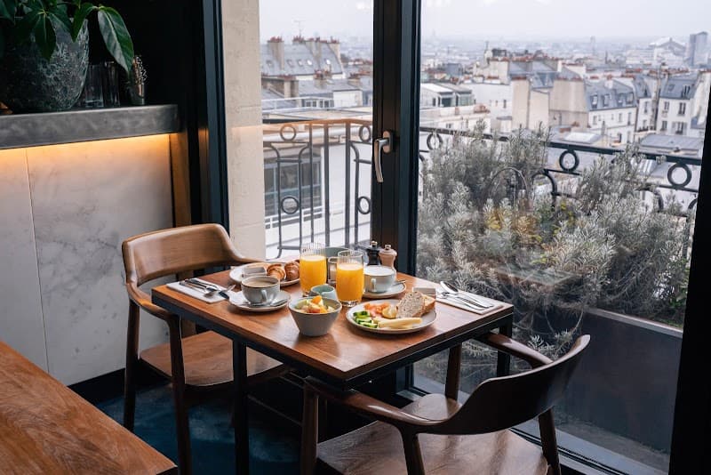 A cozy breakfast setup on a wooden table by a balcony offers a serene view of Paris rooftops, perfect for travelers seeking a charming urban retreat experience.