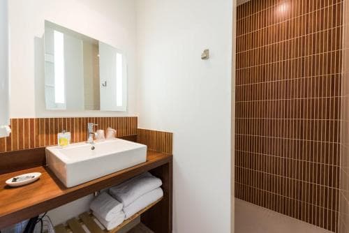 A modern bathroom features a sleek white sink on a wooden vanity, neatly folded towels, and a tiled shower wall, ideal for travelers seeking a clean and stylish accommodation space.