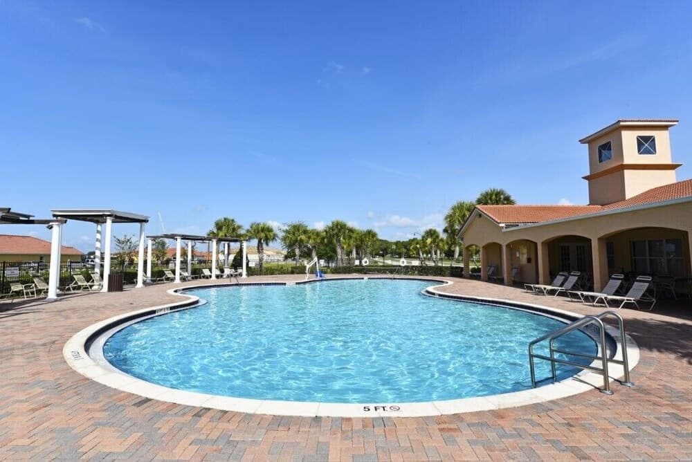 A serene resort pool area features clear blue water surrounded by lounge chairs, palm trees, and a clubhouse under a bright sky, ideal for vacation relaxation and leisure activities.