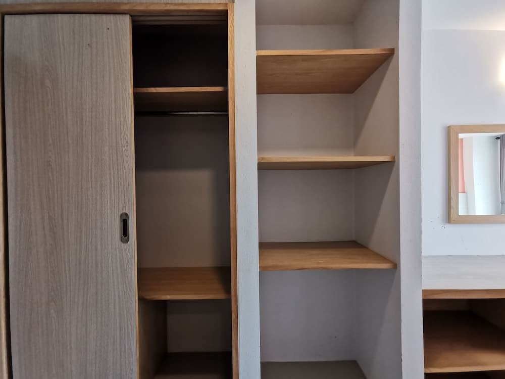 A minimalist bedroom corner features an open wooden wardrobe with empty shelves, a sleek mirror, and a clean desk, ideal for travelers seeking cozy and organized accommodation.