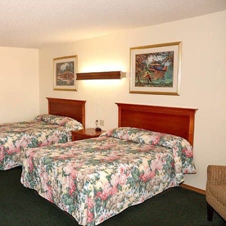 A cozy hotel room features two floral-patterned beds under warm wall sconces, framed artwork, and a wicker chair, ideal for travelers seeking comfort and relaxation.