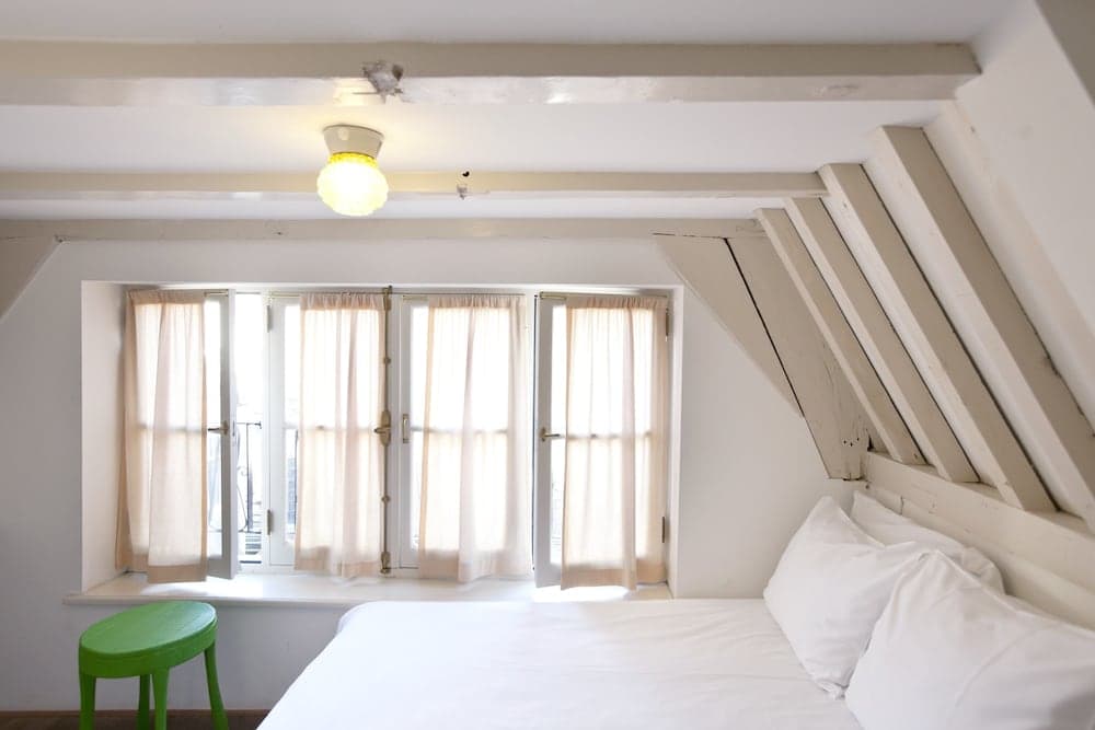 A cozy attic bedroom with sloped ceilings, wooden beams, a bright window with sheer curtains, and a green stool, ideal for travelers seeking a charming and intimate accommodation experience.
