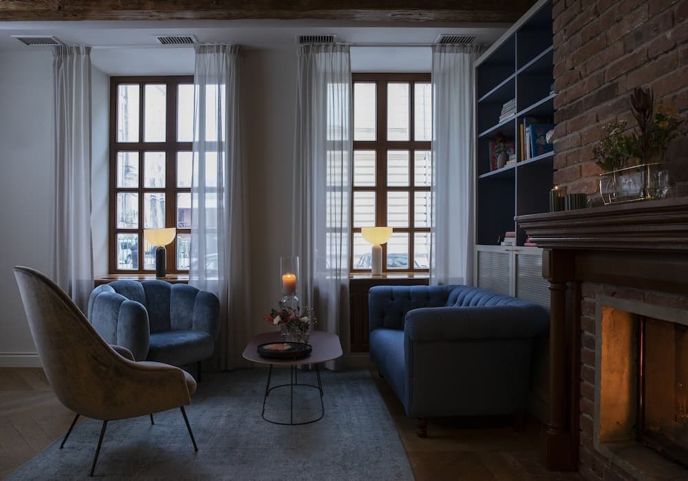 A cozy living room with soft lighting, blue velvet furniture, a brick fireplace, and large windows framed by sheer curtains creates an inviting atmosphere ideal for a relaxing getaway or home stay experience.