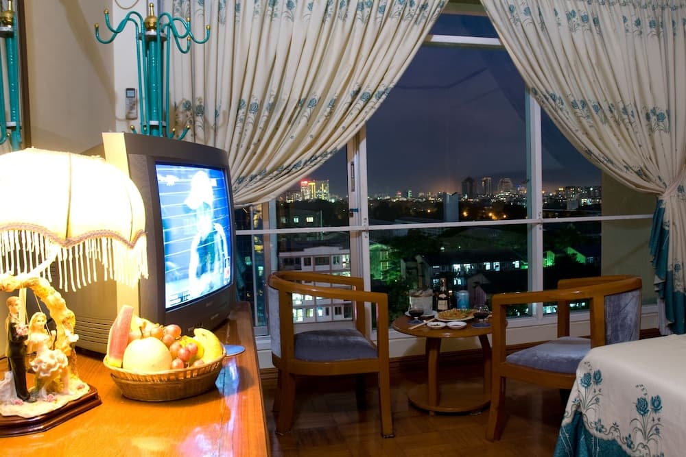 A cozy evening in a warmly lit apartment features a fruit basket on a wooden table, a vintage TV, elegant curtains framing a cityscape view, and inviting chairs set for relaxation, perfect for travelers seeking a homey urban retreat.