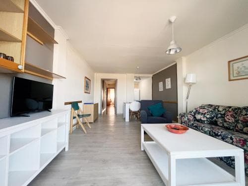 A cozy apartment interior features a modern living space with a floral-patterned sofa, sleek white furniture, and a flat-screen TV, ideal for travelers seeking comfort and convenience in a home-like setting.