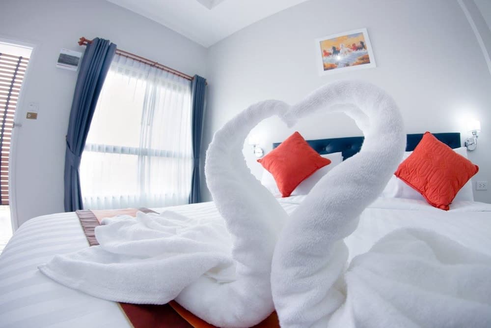 A cozy hotel room features a neatly made bed adorned with orange pillows and swan-shaped towels forming a heart, set against soft natural light from a window, ideal for romantic getaways or leisure travelers seeking comfort.