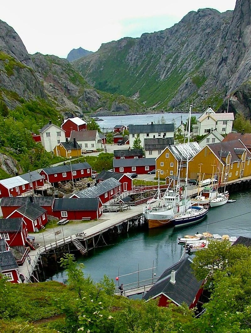 A picturesque coastal village nestled among towering mountains, featuring vibrant red and yellow houses along the waterfront, boats docked at the pier, and lush greenery, ideal for travelers seeking serene natural beauty and cultural exploration in Norway.