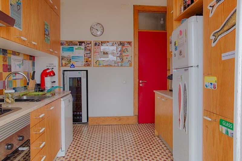 A cozy hostel kitchen in Lisbon features wooden cabinets, a tiled floor, a vibrant red door, and a fridge adorned with magnets, ideal for travelers seeking a communal cooking space.